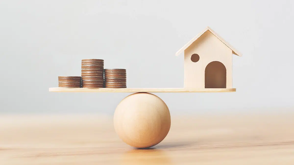 Une balance représentant l'équilibre parfait entre des pièces de monnaie et une petite maison en bois, symbolisant les aides financières pour la construction en Région Wallonne.